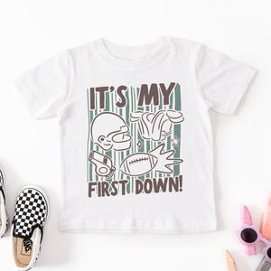May include: White t-shirt with a football-themed graphic. The design includes a football helmet, shoulder pads, a whistle, and a football, with the text "IT'S MY FIRST DOWN!" in brown and green.