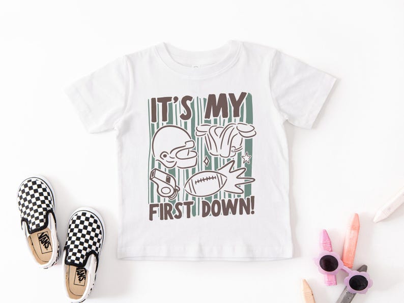May include: White t-shirt with a football-themed graphic. The design includes a football helmet, shoulder pads, a whistle, and a football, with the text "IT'S MY FIRST DOWN!" in brown and green.