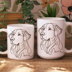May include: Two white ceramic mugs with a line drawing of a dog's face. The dog's name, "Nora", is written in cursive below the drawing. The mugs are different sizes and have white handles.