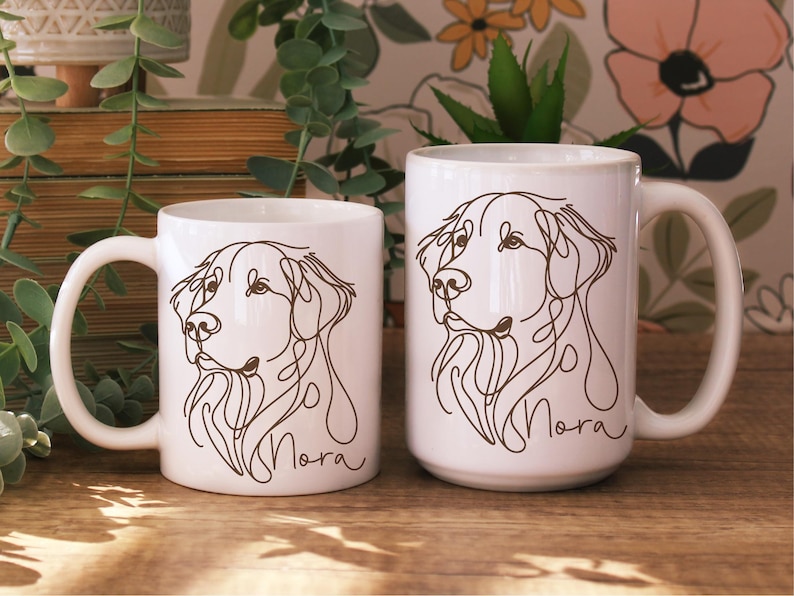 May include: Two white ceramic mugs with a line drawing of a dog's face. The dog's name, "Nora", is written in cursive below the drawing. The mugs are different sizes and have white handles.