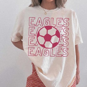 K&ouml;nnte beinhalten: Ein cremefarbenes Kurzarm-T-Shirt mit einem gro&szlig;en Grafikdruck in Dunkelrosa. Das Design besteht aus dem viermal wiederholten Wort EAGLES in einer gestapelten Umrissschrift, mit einer zentralen Fu&szlig;ball-Illustration &uuml;ber dem Text.