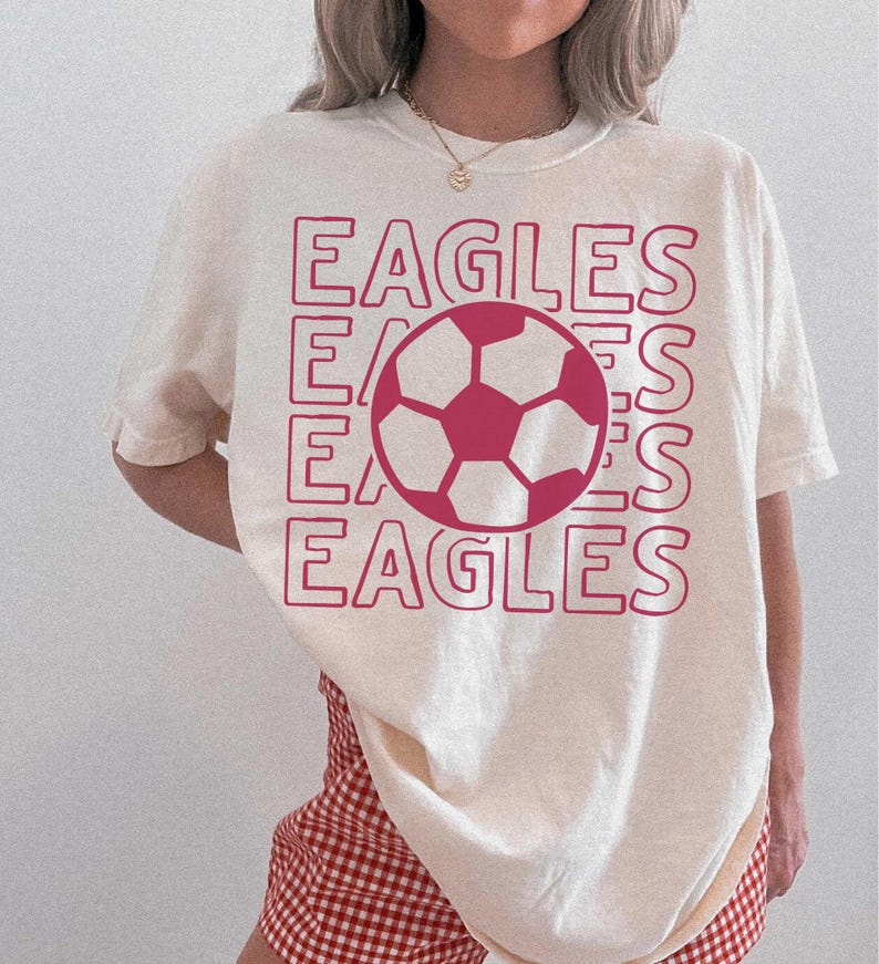 K&ouml;nnte beinhalten: Ein cremefarbenes Kurzarm-T-Shirt mit einem gro&szlig;en Grafikdruck in Dunkelrosa. Das Design besteht aus dem viermal wiederholten Wort EAGLES in einer gestapelten Umrissschrift, mit einer zentralen Fu&szlig;ball-Illustration &uuml;ber dem Text.