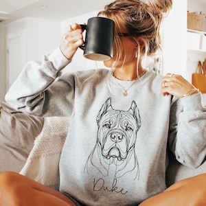 May include: A light grey sweatshirt with a line drawing of a dog's face and the name "Duke" underneath. The sweatshirt has long sleeves and a crew neck. A person is holding a black mug.