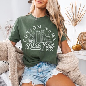 May include: Green t-shirt with a white graphic that reads "CUSTOM NAME Book Club est. 2025." The graphic includes illustrations of books, flowers, and a coffee cup. The shirt is paired with denim shorts.