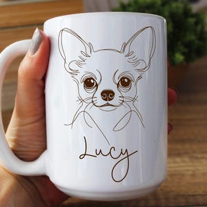 May include: White ceramic mug featuring a line drawing of a Chihuahua dog's face. The dog's name, "Lucy," is written in a cursive font below the drawing. The mug has a large handle and is held by a person.