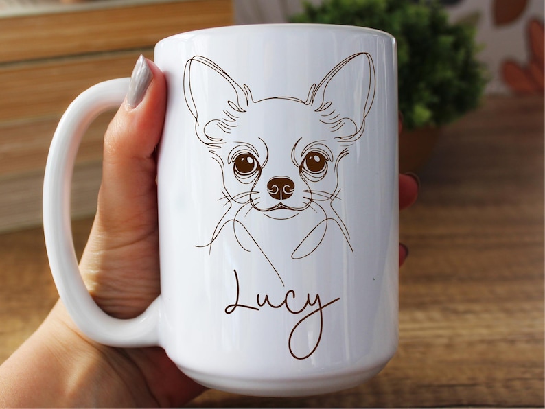 May include: White ceramic mug featuring a line drawing of a Chihuahua dog's face. The dog's name, "Lucy," is written in a cursive font below the drawing. The mug has a large handle and is held by a person.