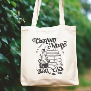 Puede incluir: Una bolsa de tela blanca con texto negro y un gr&aacute;fico. El texto dice "Custom Name Book Club est. 2024". El gr&aacute;fico es una ilustraci&oacute;n en blanco y negro de una pila de libros con una botella de poci&oacute;n y enredaderas.