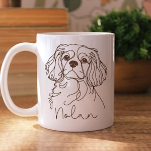 May include: White ceramic mug featuring a line drawing of a Cavalier King Charles Spaniel dog. The dog's name, "Nolan," is written in a cursive font below the drawing. The mug has a curved handle.