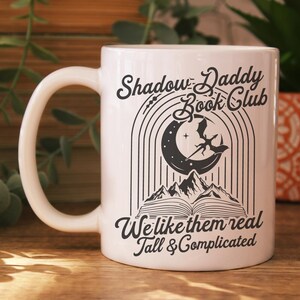 May include: White ceramic mug with a black graphic design that says "Shadow Daddy Book Club" with a crescent moon and a dragon. The text below the graphic says "We like them real tall & complicated".
