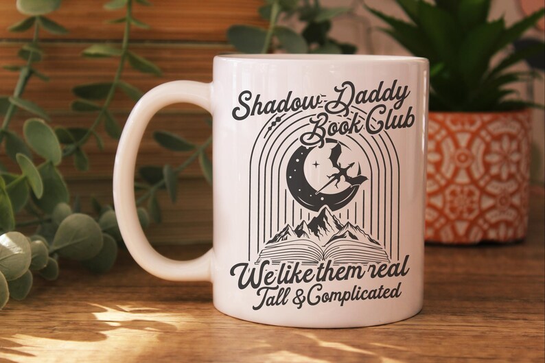 May include: White ceramic mug with a black graphic design that says "Shadow Daddy Book Club" with a crescent moon and a dragon. The text below the graphic says "We like them real tall & complicated".