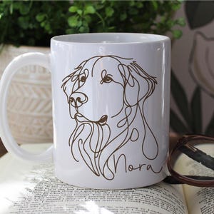 May include: White ceramic mug featuring a brown line drawing of a dog's face, with the name "Nora" in cursive script. The mug has a curved handle and is set on an open book. A pair of glasses rests on the book.