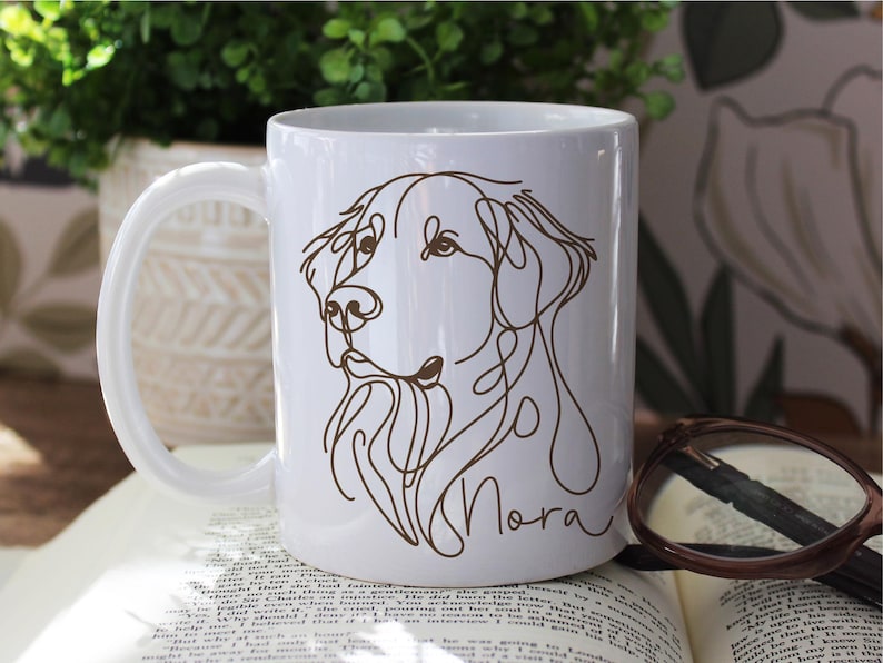May include: White ceramic mug featuring a brown line drawing of a dog's face, with the name "Nora" in cursive script. The mug has a curved handle and is set on an open book. A pair of glasses rests on the book.