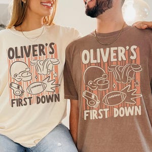 May include: Two people wearing matching beige and brown t-shirts. Each shirt has the text "OLIVER'S FIRST DOWN" with football-themed graphics including a helmet, shoulder pads, a football, and a whistle. The shirts are a relaxed fit.