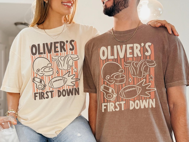 May include: Two people wearing matching beige and brown t-shirts. Each shirt has the text "OLIVER'S FIRST DOWN" with football-themed graphics including a helmet, shoulder pads, a football, and a whistle. The shirts are a relaxed fit.