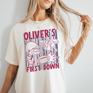 May include: Cream-colored t-shirt with a maroon and blue graphic. The graphic includes a football helmet, shoulder pads, a whistle, a football, and the text "OLIVER'S FIRST DOWN". The shirt is a casual, unisex style.