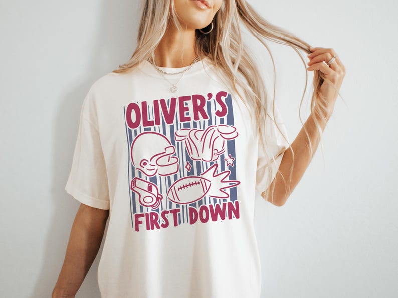 May include: Cream-colored t-shirt with a maroon and blue graphic. The graphic includes a football helmet, shoulder pads, a whistle, a football, and the text "OLIVER'S FIRST DOWN". The shirt is a casual, unisex style.