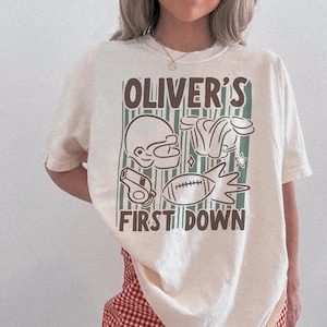 May include: Cream-colored t-shirt with a football-themed graphic. The design includes a football helmet, shoulder pads, a whistle, and a football, with the text "OLIVER'S FIRST DOWN" in brown. The shirt is a casual, unisex style.