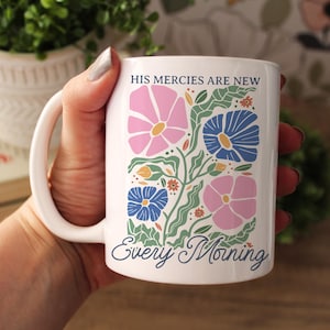 K&ouml;nnte beinhalten: Wei&szlig;e Keramik-Tasse mit einem floralen Design in Rosa und Blau, mit dem Text "HIS mercies are new Every Morning". Die Tasse hat einen wei&szlig;en Henkel und wird von einer Person gehalten.