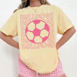 K&ouml;nnte beinhalten: Ein hellgelbes Kurzarm-T-Shirt mit einer pinkfarbenen Fu&szlig;ballgrafik in der Mitte der Brust. Das Wort BULLDOGS ist in einer sich wiederholenden, gestapelten pinkfarbenen Umriss-Schrift hinter der Fu&szlig;ballgrafik gedruckt, was einen Schichteffekt erzeugt.