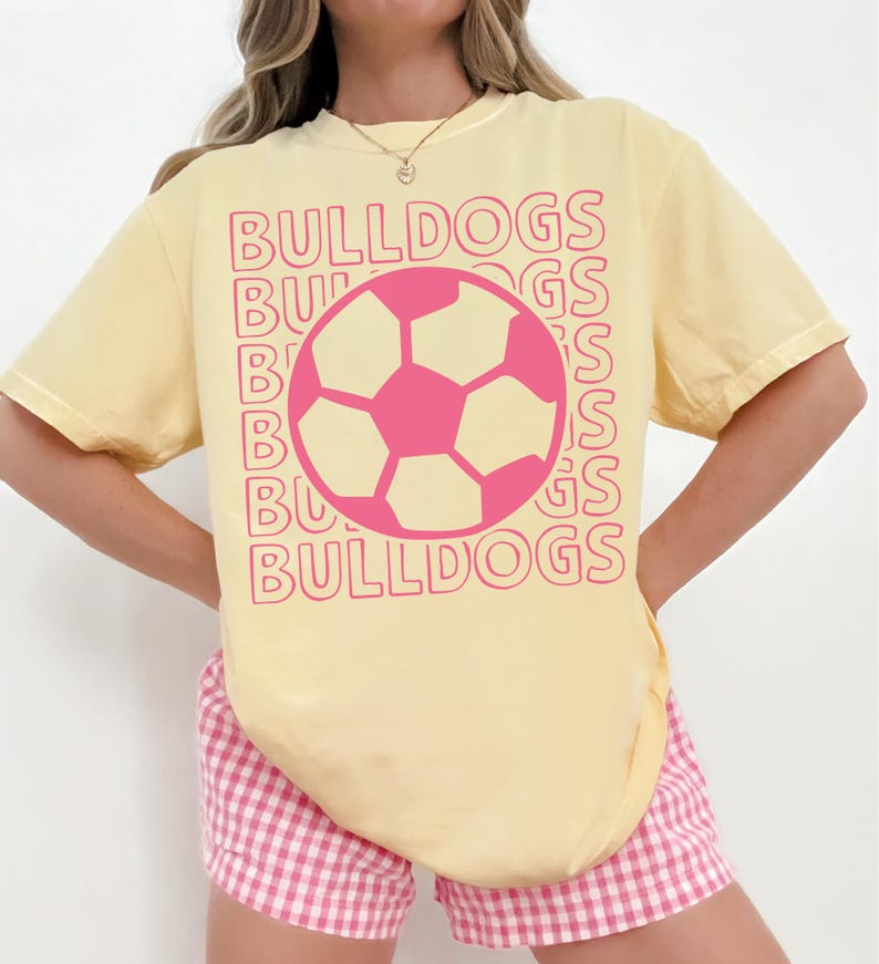 K&ouml;nnte beinhalten: Ein hellgelbes Kurzarm-T-Shirt mit einer pinkfarbenen Fu&szlig;ballgrafik in der Mitte der Brust. Das Wort BULLDOGS ist in einer sich wiederholenden, gestapelten pinkfarbenen Umriss-Schrift hinter der Fu&szlig;ballgrafik gedruckt, was einen Schichteffekt erzeugt.