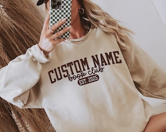 May include: A beige sweatshirt with the text "CUSTOM NAME book club EST. 2025" in dark brown lettering. The sweatshirt has long sleeves and a crew neck. The image shows a person holding a phone.