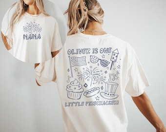 Puede incluir: Camiseta color crema con gráficos azul marino. La parte delantera presenta "MAMA" con fuegos artificiales. La parte trasera muestra "OLIVER IS ONE" con ilustraciones patrióticas, incluyendo una bandera, fuegos artificiales y una magdalena. También está el texto "LITTLE FIRECRACKER!"