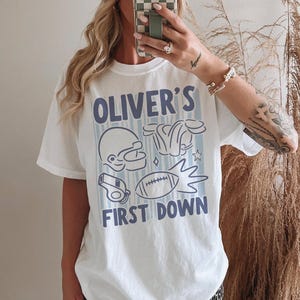 May include: White t-shirt with a blue and white football-themed graphic. The design includes a football helmet, shoulder pads, a whistle, and a football. Text on the shirt reads "OLIVER'S FIRST DOWN."