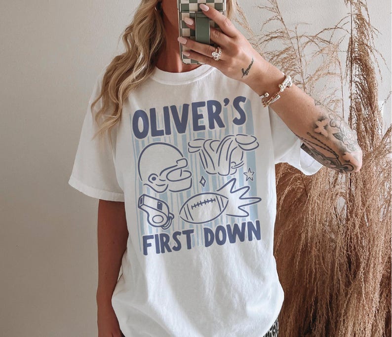 May include: White t-shirt with a blue and white football-themed graphic. The design includes a football helmet, shoulder pads, a whistle, and a football. Text on the shirt reads "OLIVER'S FIRST DOWN."