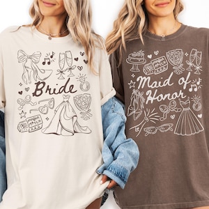 May include: Two t-shirts, one cream-colored and one brown, featuring hand-drawn illustrations. The cream shirt says "Bride" with wedding-themed doodles. The brown shirt says "Maid of Honor" with similar illustrations. Both shirts have short sleeves.