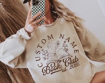 May include: Beige sweatshirt with a brown graphic design. The design features an open book with floral elements and the text "CUSTOM NAME Book Club est. 2025". The sweatshirt has long sleeves and a crew neck.
