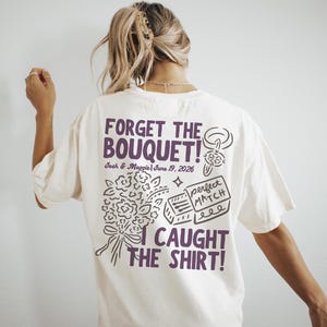 May include: Cream-colored t-shirt with the text "FORGET THE BOUQUET! I CAUGHT THE SHIRT!" in purple. The shirt also features a bouquet illustration, a ring, and a matchbox design. The date "June 19, 2026" is also printed on the shirt.
