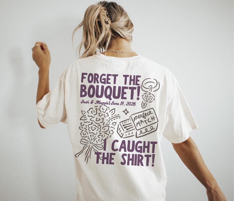 May include: Cream-colored t-shirt with the text "FORGET THE BOUQUET! I CAUGHT THE SHIRT!" in purple. The shirt also features a bouquet illustration, a ring, and a matchbox design. The date "June 19, 2026" is also printed on the shirt.