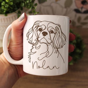 May include: White ceramic mug featuring a line drawing of a Cavalier King Charles Spaniel dog. The dog's name, "Nolan", is written in a cursive font below the illustration. The mug has a white handle.