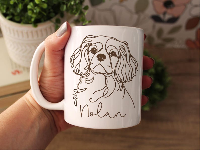 May include: White ceramic mug featuring a line drawing of a Cavalier King Charles Spaniel dog. The dog's name, "Nolan", is written in a cursive font below the illustration. The mug has a white handle.