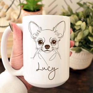 May include: White ceramic mug featuring a line drawing of a Chihuahua's face. The dog's features are detailed with brown lines, and the name "Lucy" is written below. The mug has a large handle and is held by a person.