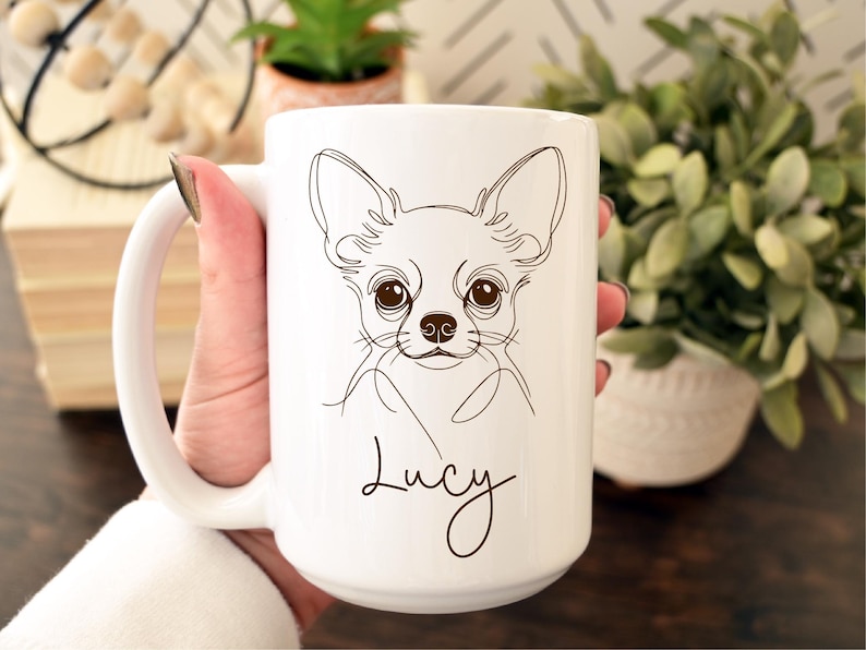 May include: White ceramic mug featuring a line drawing of a Chihuahua's face. The dog's features are detailed with brown lines, and the name "Lucy" is written below. The mug has a large handle and is held by a person.