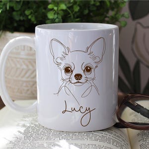 May include: White ceramic mug featuring a line drawing of a Chihuahua dog with large brown eyes and ears. The name "Lucy" is written in a cursive font below the dog's face. The mug has a curved handle.