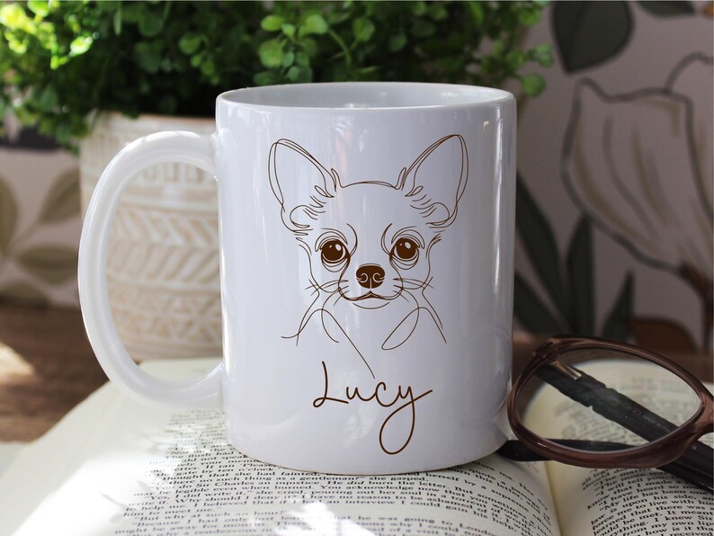 May include: White ceramic mug featuring a line drawing of a Chihuahua dog with large brown eyes and ears. The name "Lucy" is written in a cursive font below the dog's face. The mug has a curved handle.