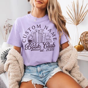 May include: A light purple t-shirt with a circular design that reads "CUSTOM NAME Book Club est. 2025". The design includes illustrations of books, flowers, and a coffee cup. The shirt is worn with denim shorts and a cream-colored cardigan.