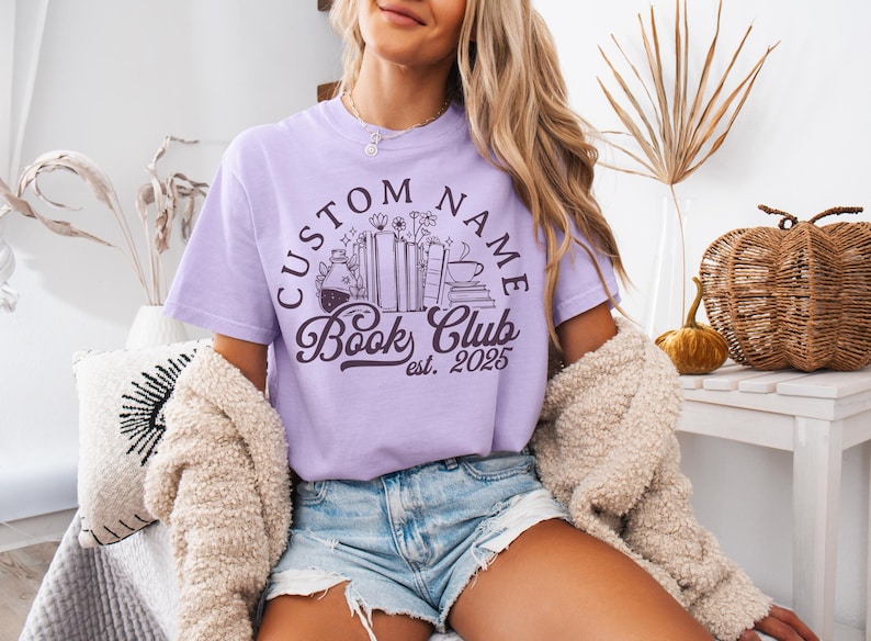 May include: A light purple t-shirt with a circular design that reads "CUSTOM NAME Book Club est. 2025". The design includes illustrations of books, flowers, and a coffee cup. The shirt is worn with denim shorts and a cream-colored cardigan.