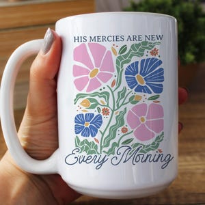 K&ouml;nnte beinhalten: Wei&szlig;er Keramikbecher mit einem Blumendesign in Rosa, Blau und Gr&uuml;n. Der Becher tr&auml;gt den Text "HIS MERCIES ARE NEW Every Morning" in einer dekorativen Schriftart. Der Becher hat einen gro&szlig;en Henkel.