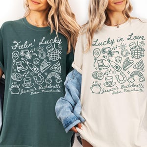 May include: Two t-shirts, one dark green and one cream, with St. Patrick's Day themed graphics. The green shirt says "Feelin' Lucky" and the cream shirt says "Lucky in Love." Both shirts feature illustrations of shamrocks, rainbows, and other festive icons. The text "Jessica's Bachelorette, Boston, Massachusetts" is also printed on both shirts.
