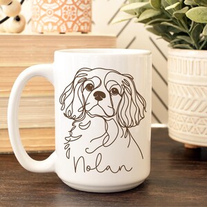 May include: White ceramic mug featuring a line drawing of a Cavalier King Charles Spaniel dog. The dog's name, "Nolan," is written in a cursive font below the illustration. The mug has a large handle and is suitable for hot beverages.
