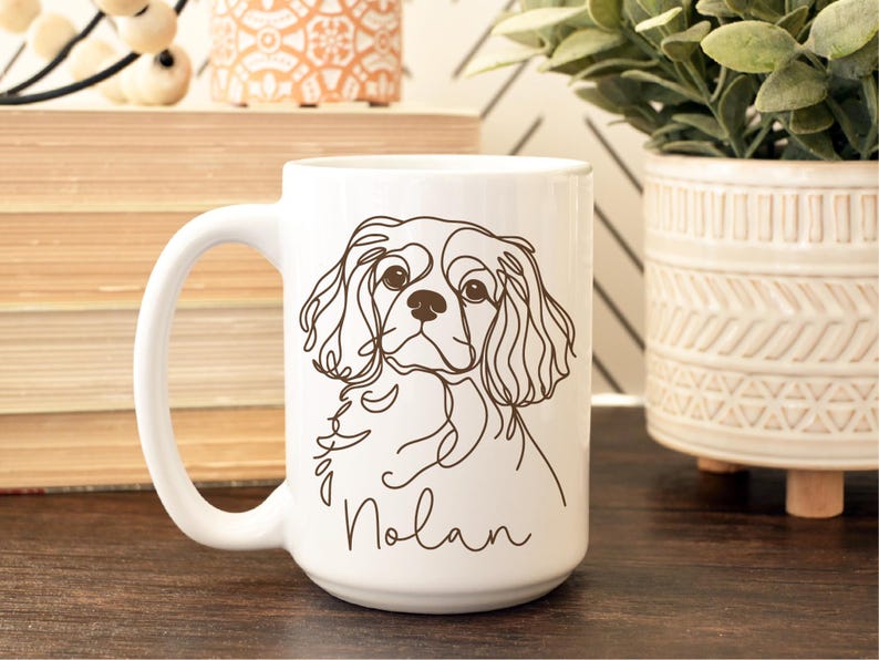 May include: White ceramic mug featuring a line drawing of a Cavalier King Charles Spaniel dog. The dog's name, "Nolan," is written in a cursive font below the illustration. The mug has a large handle and is suitable for hot beverages.