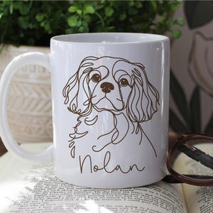 May include: White ceramic mug featuring a brown line drawing of a Cavalier King Charles Spaniel dog. The dog's name, "Nolan," is written in a script font below the illustration. The mug has a curved handle.