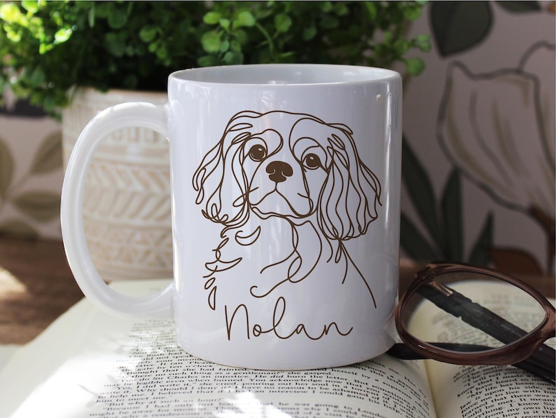 May include: White ceramic mug featuring a brown line drawing of a Cavalier King Charles Spaniel dog. The dog's name, "Nolan," is written in a script font below the illustration. The mug has a curved handle.