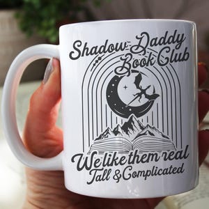 May include: White ceramic mug with a black and white design. The design features a crescent moon with a dragon flying above it, a book, and the text "Shadow Daddy Book Club We like them real Tall & Complicated".