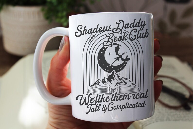 May include: White ceramic mug with a black and white design. The design features a crescent moon with a dragon flying above it, a book, and the text "Shadow Daddy Book Club We like them real Tall & Complicated".