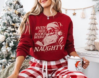May include: A red sweatshirt with the text "EVEN HER BOOKS WERE NAUGHTY" and a Santa Claus graphic. The person is wearing red and white plaid pajama pants and holding a mug with a Santa Claus design.
