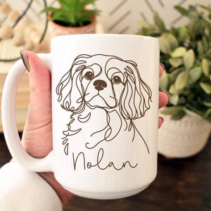 May include: White ceramic mug featuring a brown line drawing of a Cavalier King Charles Spaniel dog. The dog's name, "Nolan," is written in a script font below the dog's image. The mug has a large handle.